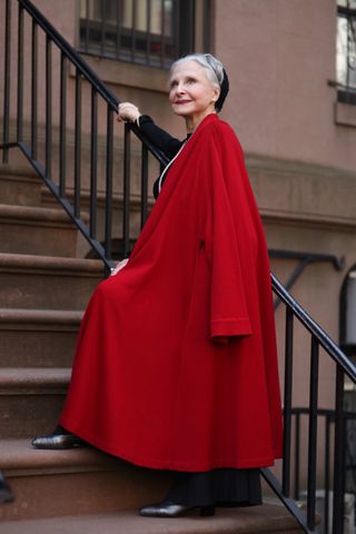 Joyce Carpati in an over 60 outfit featuring a bright red cape, black dress, boots, and pearls.