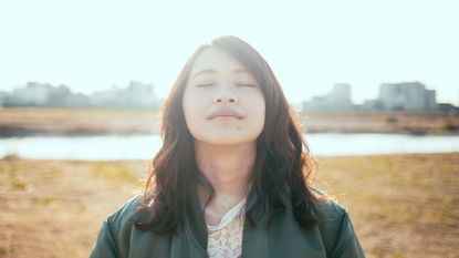 A woman stands in a field in the sunshine and takes a deep breath.