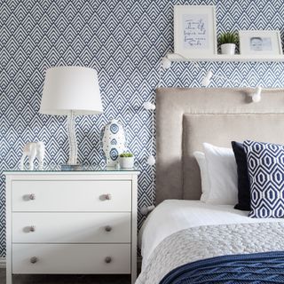 Bed with grey headboard with white bedding and patterned cushions and throw, in front of blue and white patterned wallpaper
