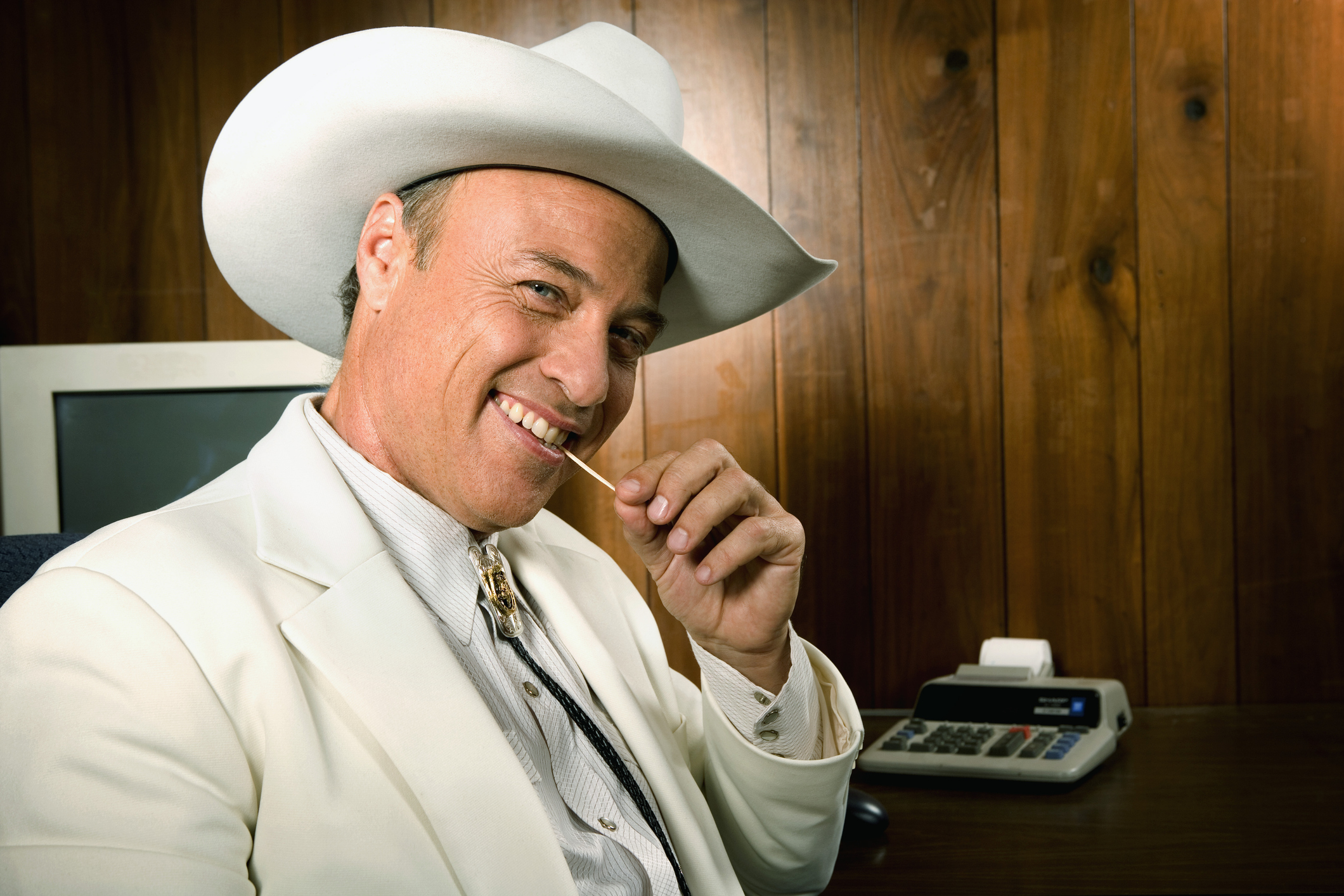 A sly looking man in a cowboy hat with a toothpick in his mouth smiles at the camera. He appears to be untrustworthy.