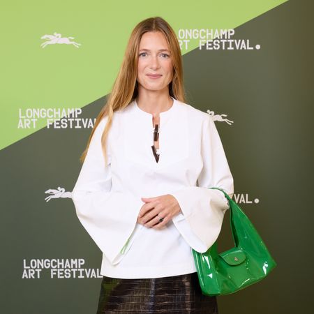 Alizee Thevenet in a white top and leather skirt standing in front of a green Longchamp step and repeat
