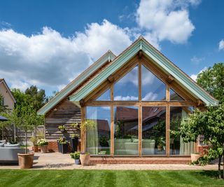 A small single storey detached oak frame home. The roof is pitched and it has a completely glazed frontage. The lawn in front is neat and there is a well-kept patio to the left of the house