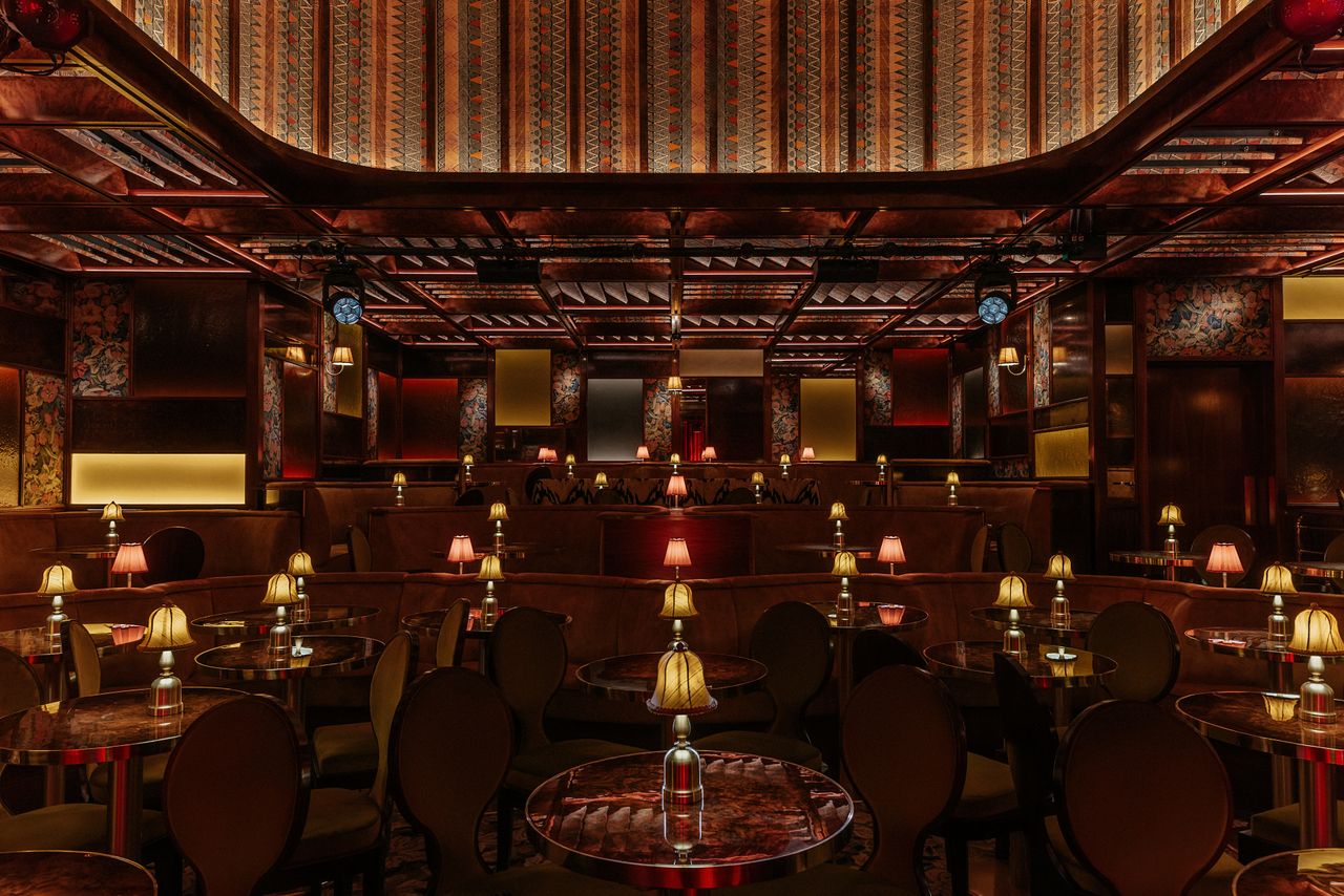 A dimly lit, textiles and lacquered wood-clad jazz club with lots of softly glowing table lamps, performers playing live, and a red-tinted light.
