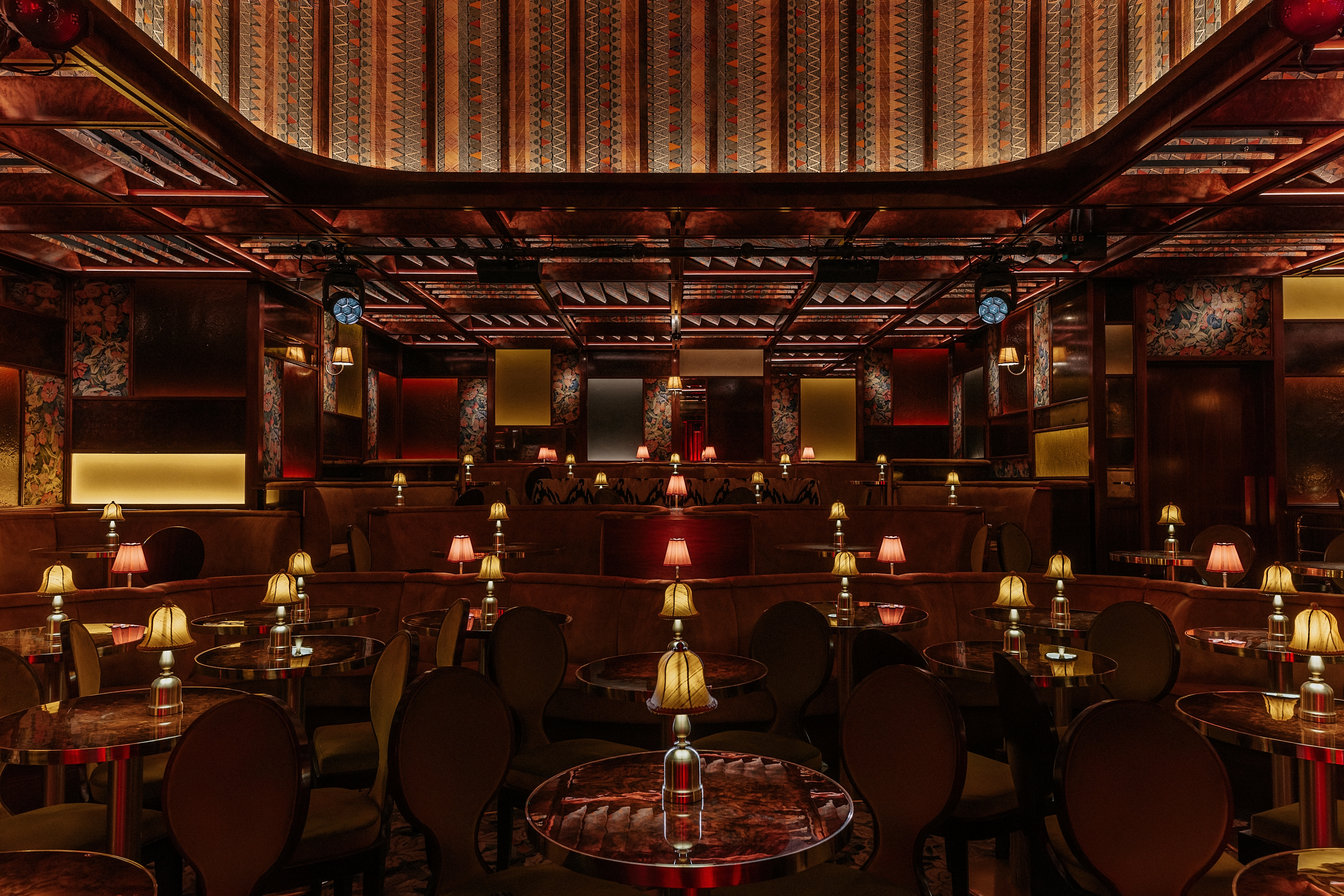 A dimly lit, textiles and lacquered wood-clad jazz club with lots of softly glowing table lamps, performers playing live, and a red-tinted light.