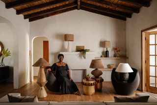 A woman in a black dress poses gracefully sitting on a minimalist armchair in black in a room beautifully decorated with rattan, wood, and glass lamps and homeware.