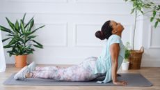 woman on a grey exercise mat on her front lifting her chest up in a spinal extension. there are two indoor plants behind her.