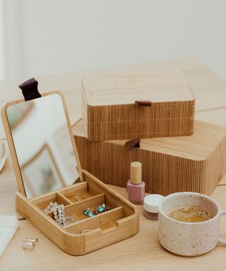 A light wooden table with a wooden jewelry box with jewelry in it, two stacked wooden boxes on top of each other, a bottle of pink nail varnish, and a white coffee cup
