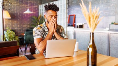 A man looks upset as he looks at his laptop at his kitchen table.