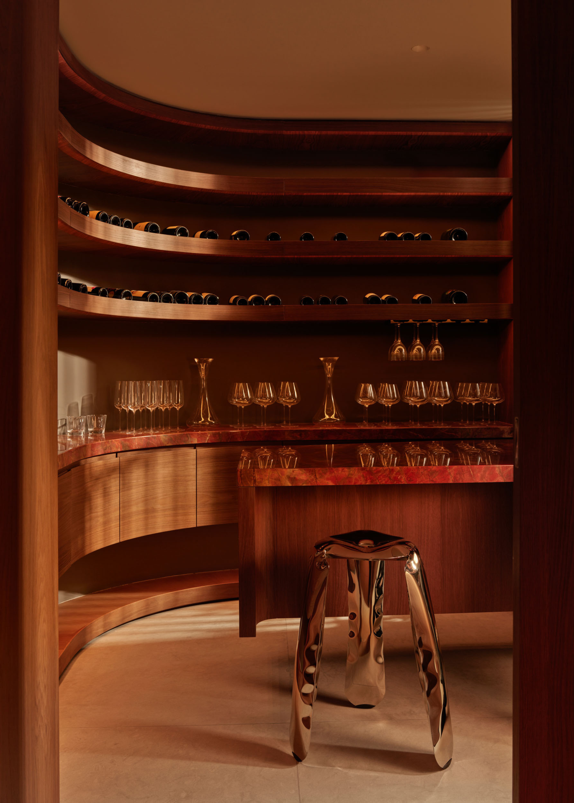 A curved bar room with red stone countertops, wooden cabinets, a steel deflated-style stool, rows of wine glasses, bottles, and champagne coupes by decanters