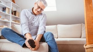 older man wearing button up, jeans and socks sits on a couch holding his right foot, as if in pain