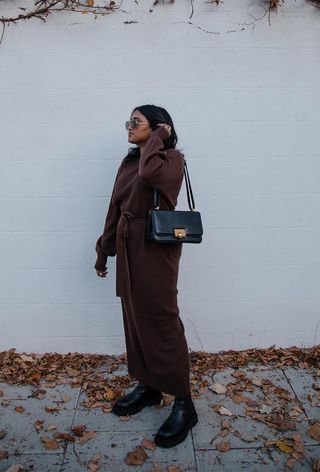 how to wear Chelsea boots is shown in a photo of a woman standing outside wearing a brown knit sweater dress with black chelsea boots and a black shoulder bag