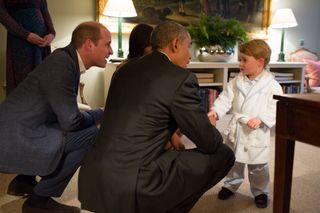 Prince William and Barack Obama kneeling down on the carpet talking to Prince George, who is wearing a white bathrobe and checked pajamas