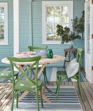 Porch/patio with duck egg blue painted shiplap, round table with green painted chairs, stripe blue rug and cushions, tableware, vase of foliage on table