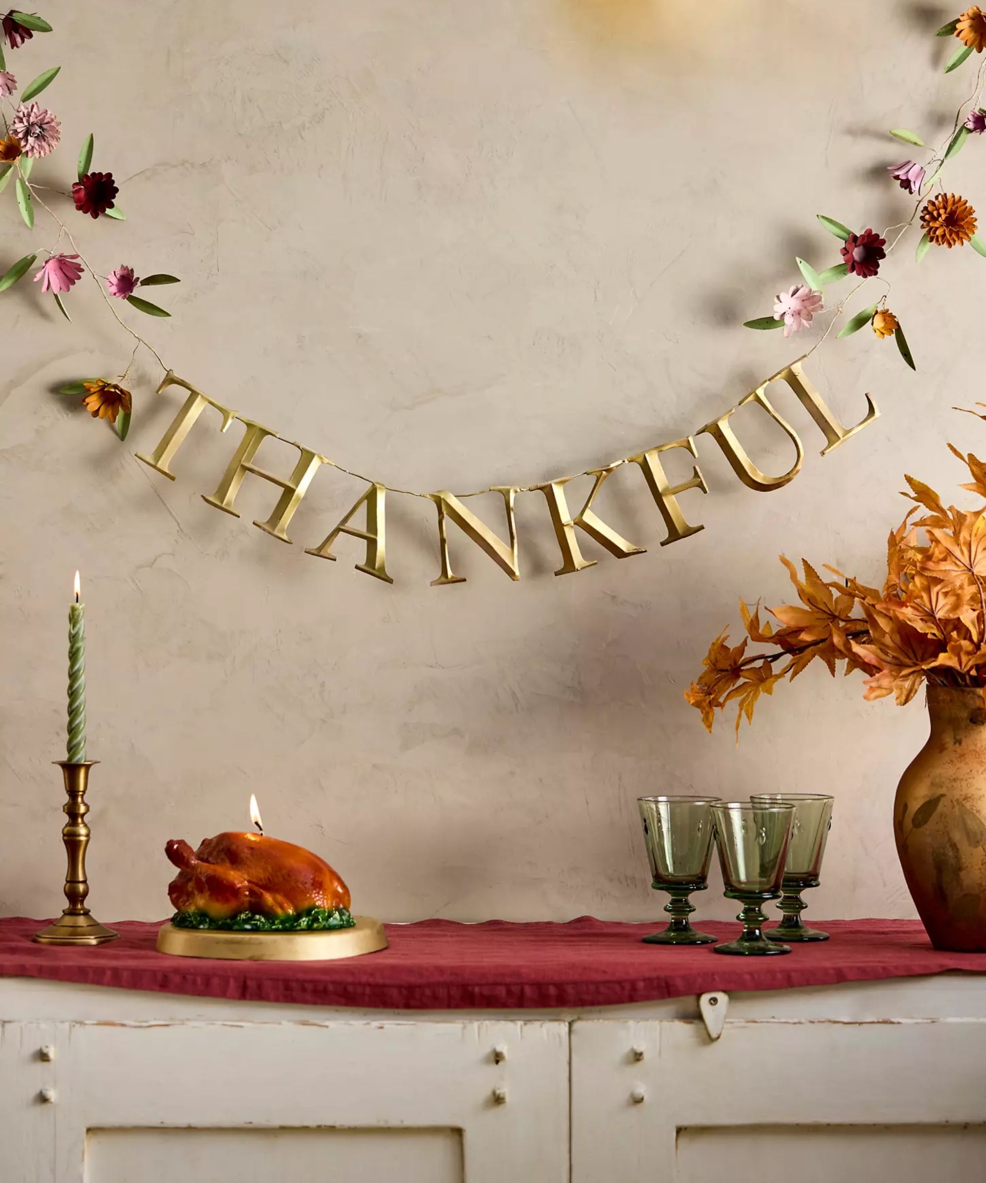 rustic white wooden buffet table styled with a red linen tablecloth, green wine glasses, a turkey shaped candle and a vase with a &#039;thankful&#039; garland hung above