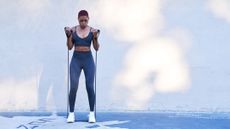 A woman stands with a resistance band underneath her feet and the ends of the bands in her hands. Her elbows are bent and her hands are near her shoulders. She is exhaling forcefully as she executes the move.