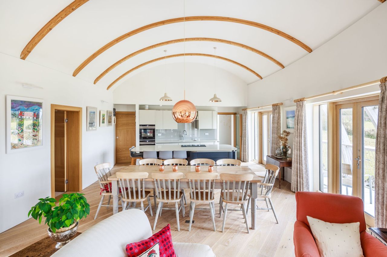 A kitchen dining room with a curved ceiling with timber beams and a kitchen island in front of a dining room table