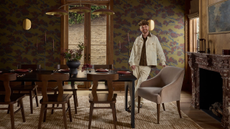 a dining room with a large dining table, several wood dining chairs, a brown rug, floral wallpaper, and a man standing in the room