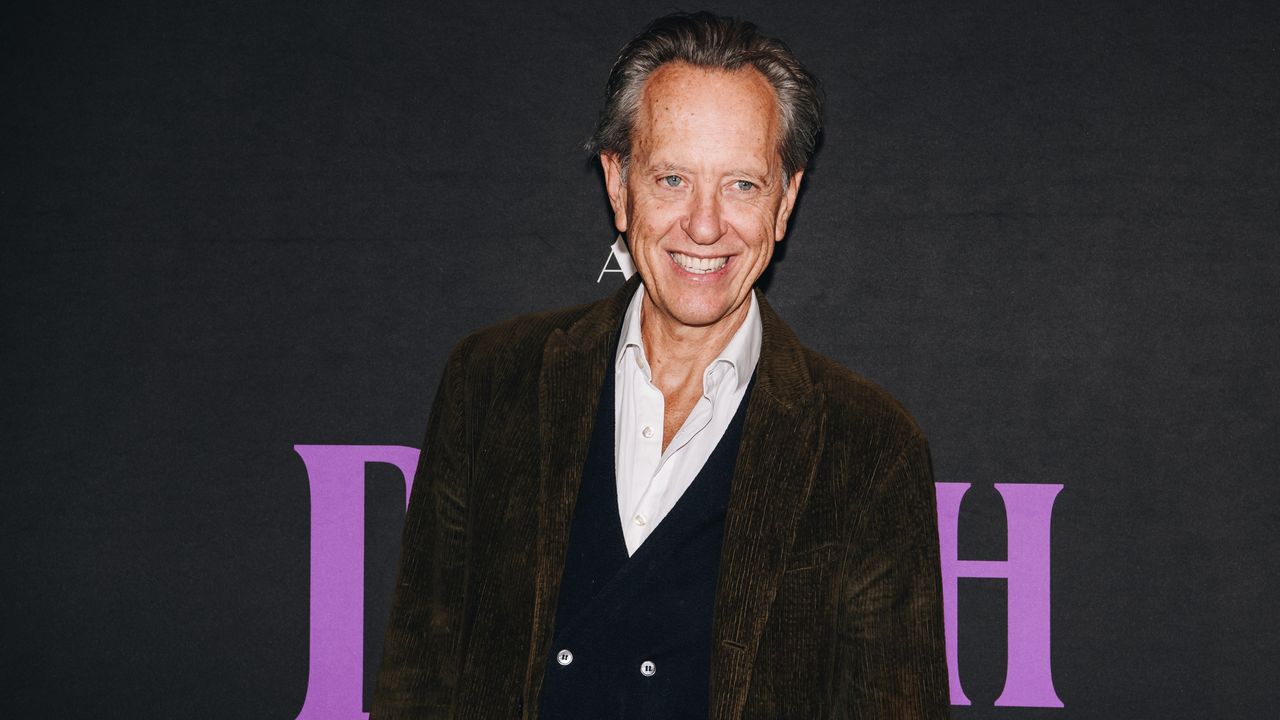 Image of Richard Grant in a brown blazer and white button-down with a black waistcoat, standing in front of a black background with purple text.