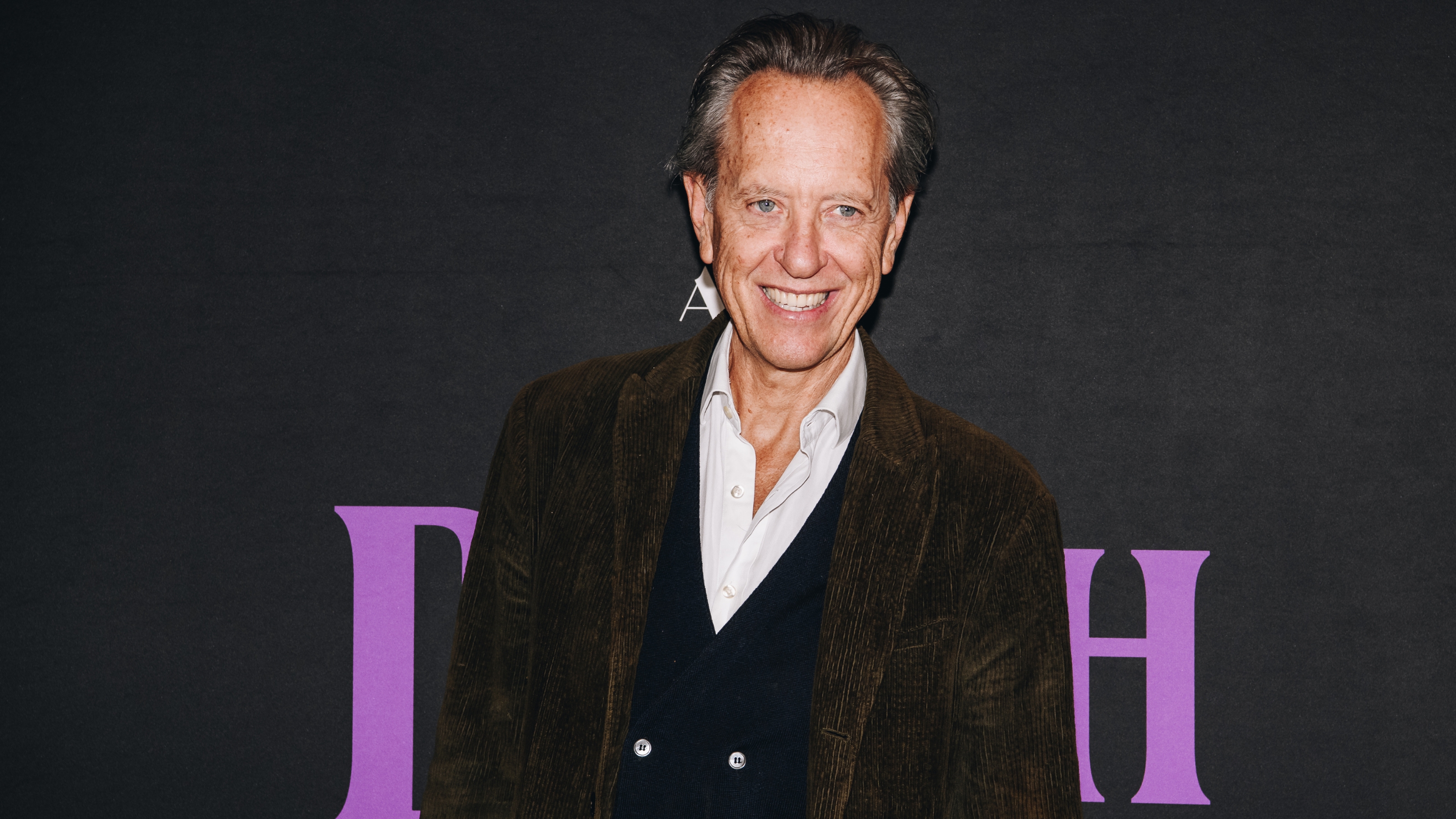 Image of Richard Grant in a brown blazer and white button-down with a black waistcoat, standing in front of a black background with purple text.