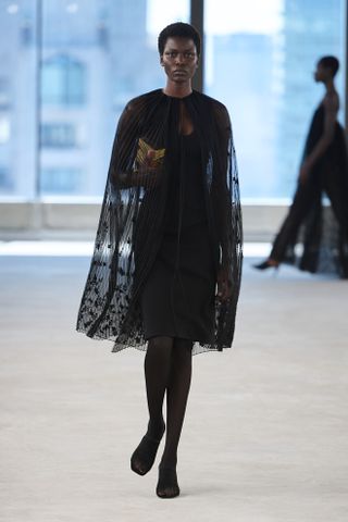 A model in Toteme Spring 2025 wears a black sheer cape and black dress on the runway