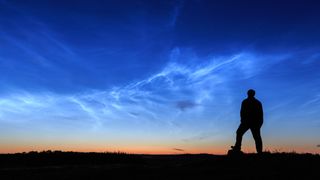 Silhouette of a person looking at the sky that contains thin wispy clouds known as noctilucent clouds. 