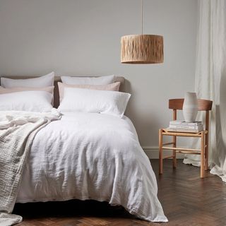 A bedroom with a low hanging light raffia pendant and a bed dressed in white linen bedlinen with a wooden chair holding books and a vase