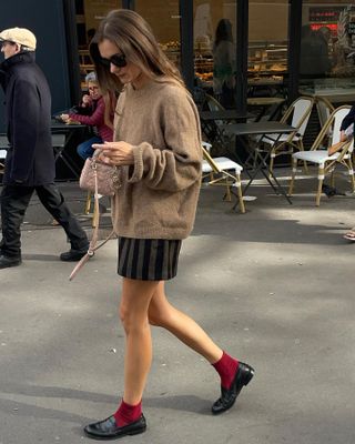 French influencer @annelauremais wearing a camel crewneck sweater, striped mini skirt, mini Dior Lady Bag, red crew socks, and black loafers