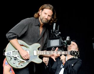 Bradley Cooper plays guitar on stage as he is filmed by a camera operator behind him.