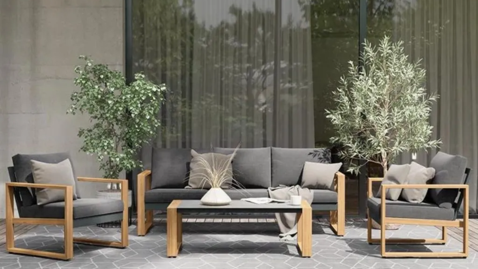 an outdoor patio with a couch and two accent chairs with gray pillows, a coffee table, a large gray rug, and decorative plants in the background