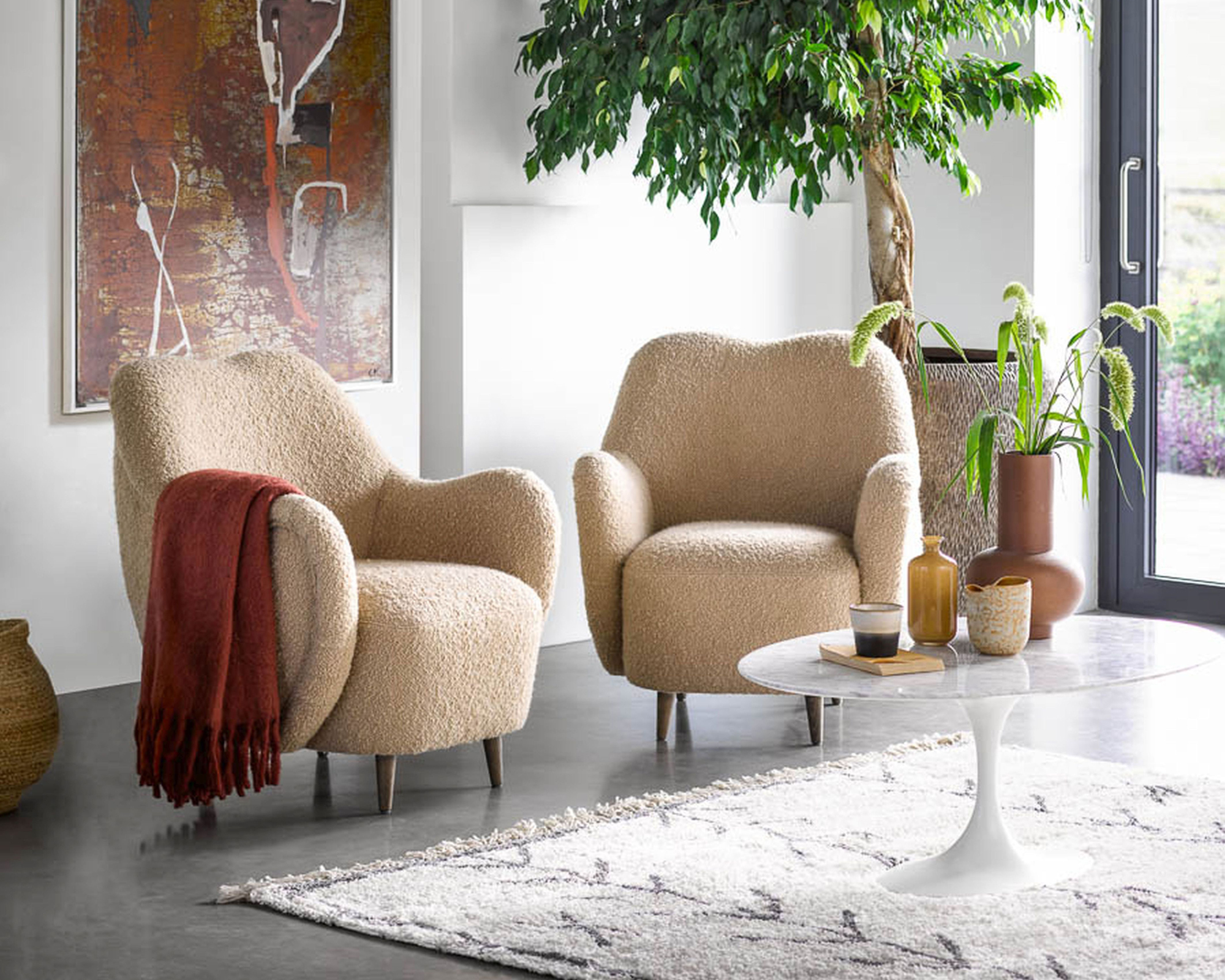 A living room with a pair of curved beige boucle armchairs with modern art on the wall and indoor tree