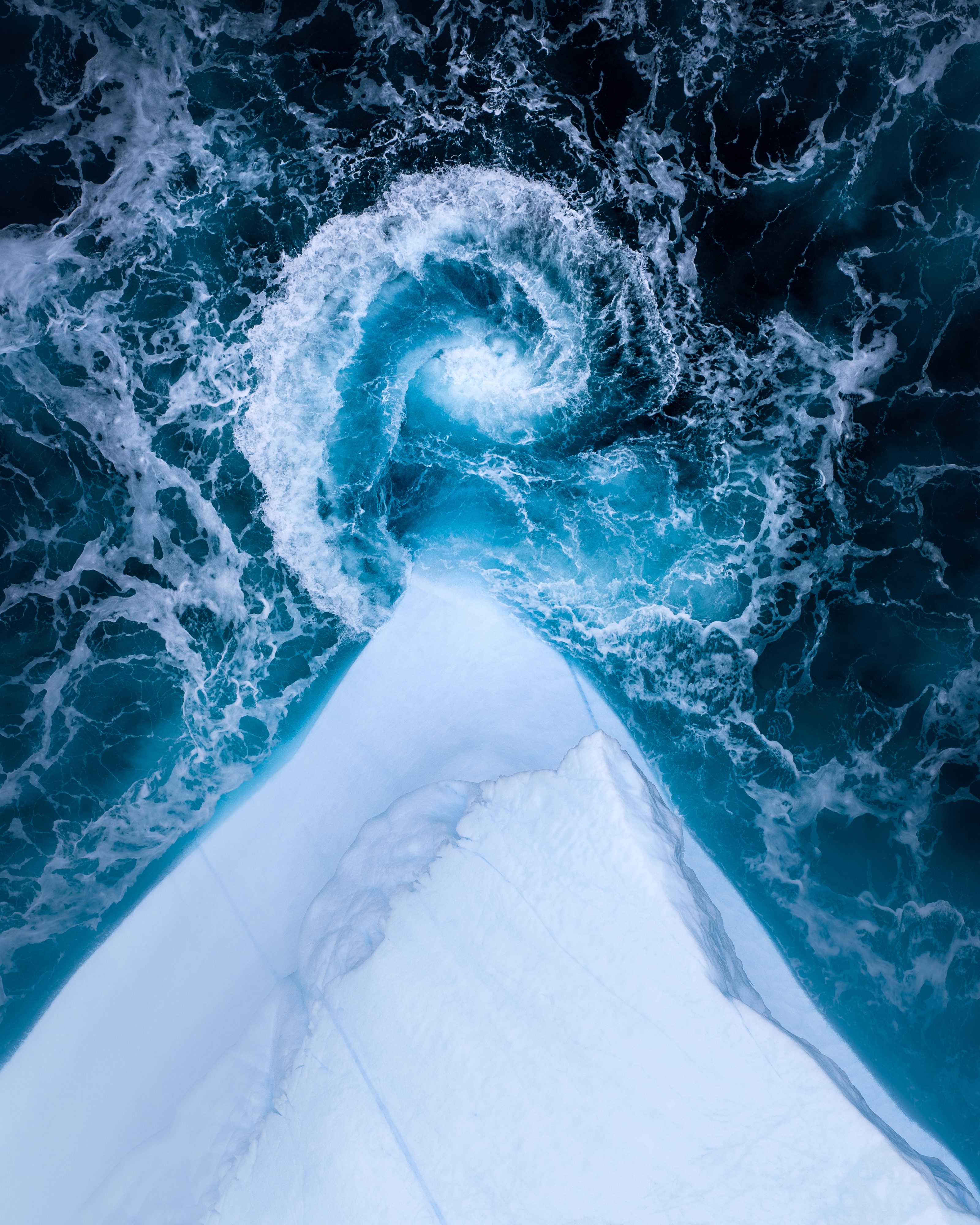 An aerial view shows the sharp, icy white edge of an iceberg meeting dark blue ocean water, where a foamy, blue-tinged wave curls into a perfect spiral