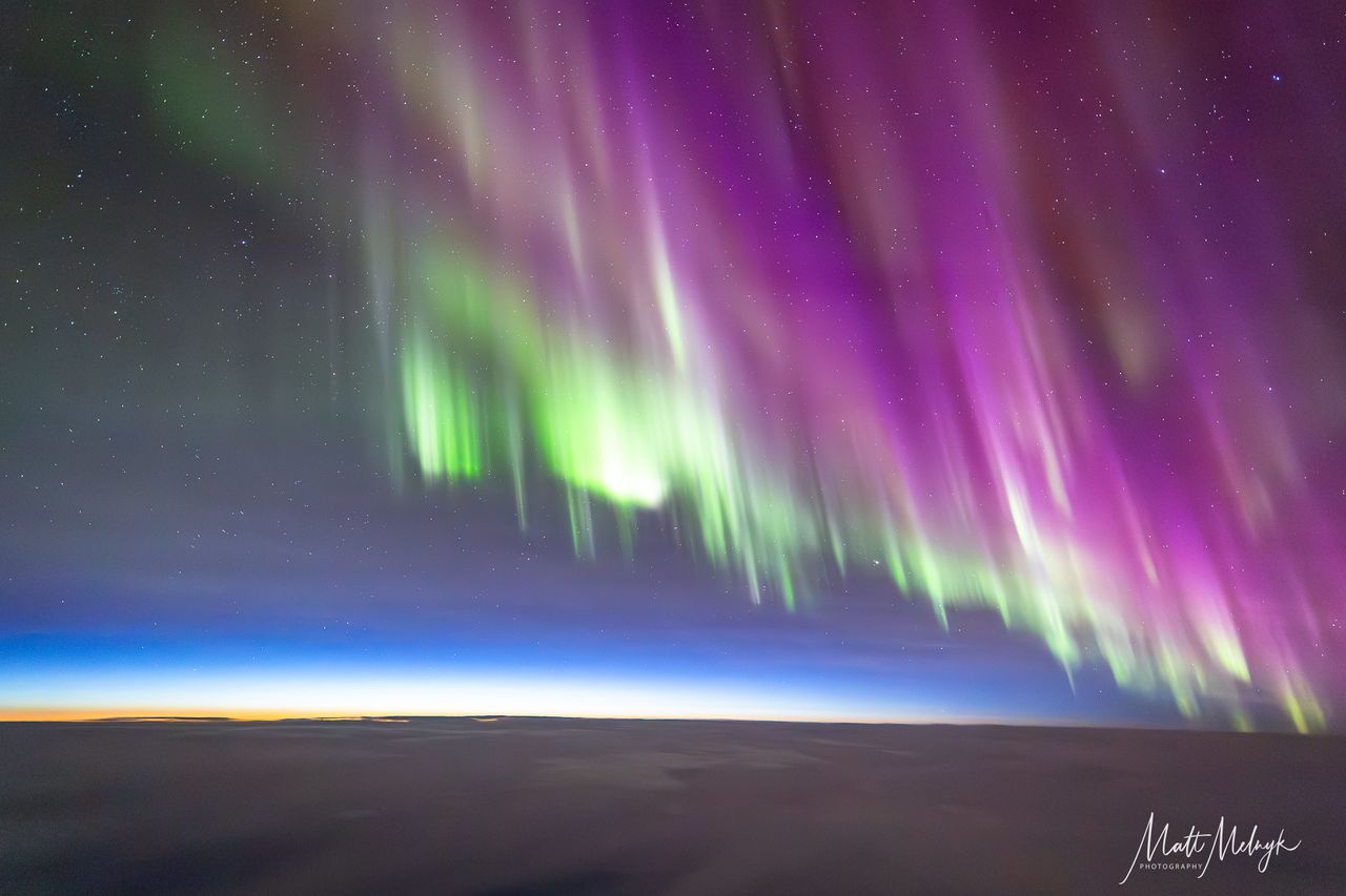 curtains of magenta and green light dance in the sky, a thin blue line marking Earth's atmosphere is visible in the distance.
