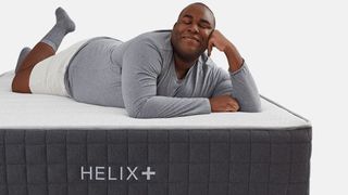 A man dressed in a gray lounge top and white lounge shorts lies on top of the Helix+ mattress, smiling