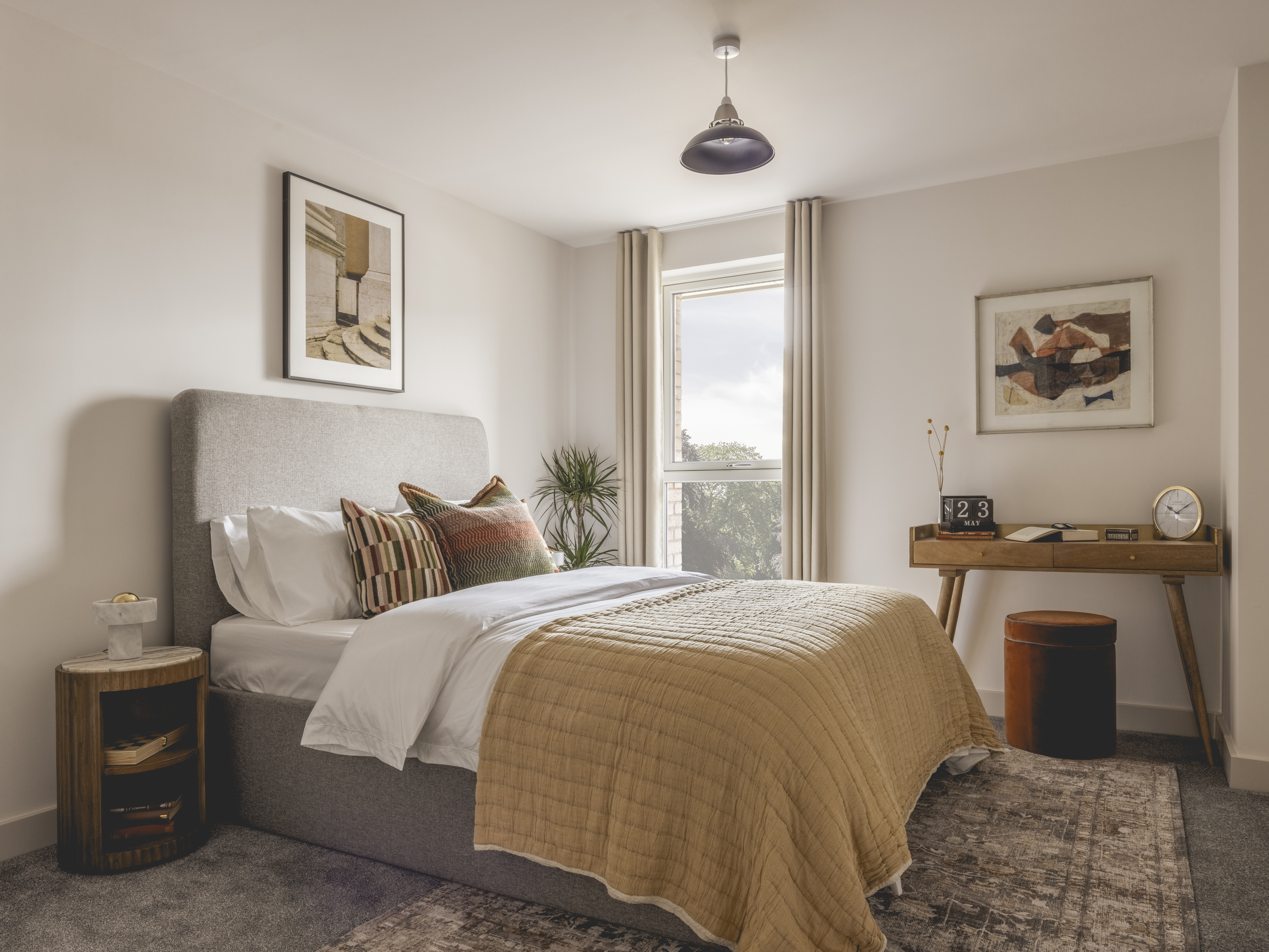A cozily decorated bedroom boasts a grey-toned rug with matching headboard, softly looking bedding topped with a yellow quilt, and colorful abstract artwork alongside wooden furniture.