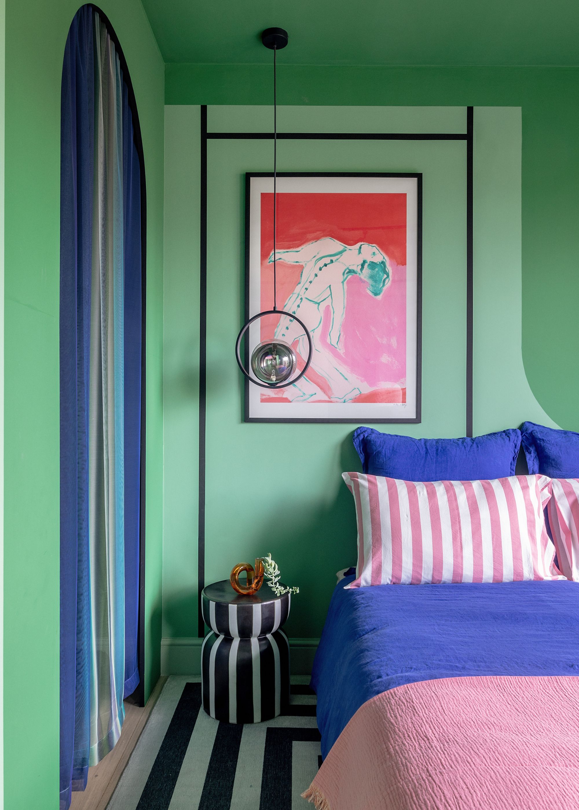 A bedroom with various shades of green paint on the walls and ceiling, bright pink artwork, a black and white nightstand and rug, and a bed with electric blue and pink bedding.