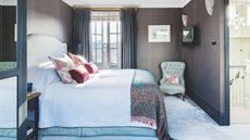 Bedroom with modern dark browny grey textured wallpaper and midnight blue curtains and door frame. In the background is a light blue period chair and accent pink cushions.