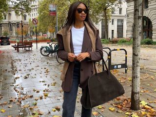 Woman wears grey jeans, a white t-shirt, a brown blazer with a sweater draped over it and black pointy-toe boots. She's carrying a The Row Margaux bag.