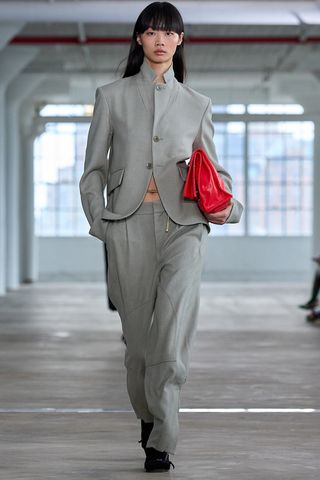 how to wear a suit is shown in a photo of a model on the runway of Tibi's spring 2025 runway show, wearing a gray blazer with the collar popped, styled with matching baggy gray trousers, a gold chain belt, a red clutch bag, black socks, and black strappy sandals