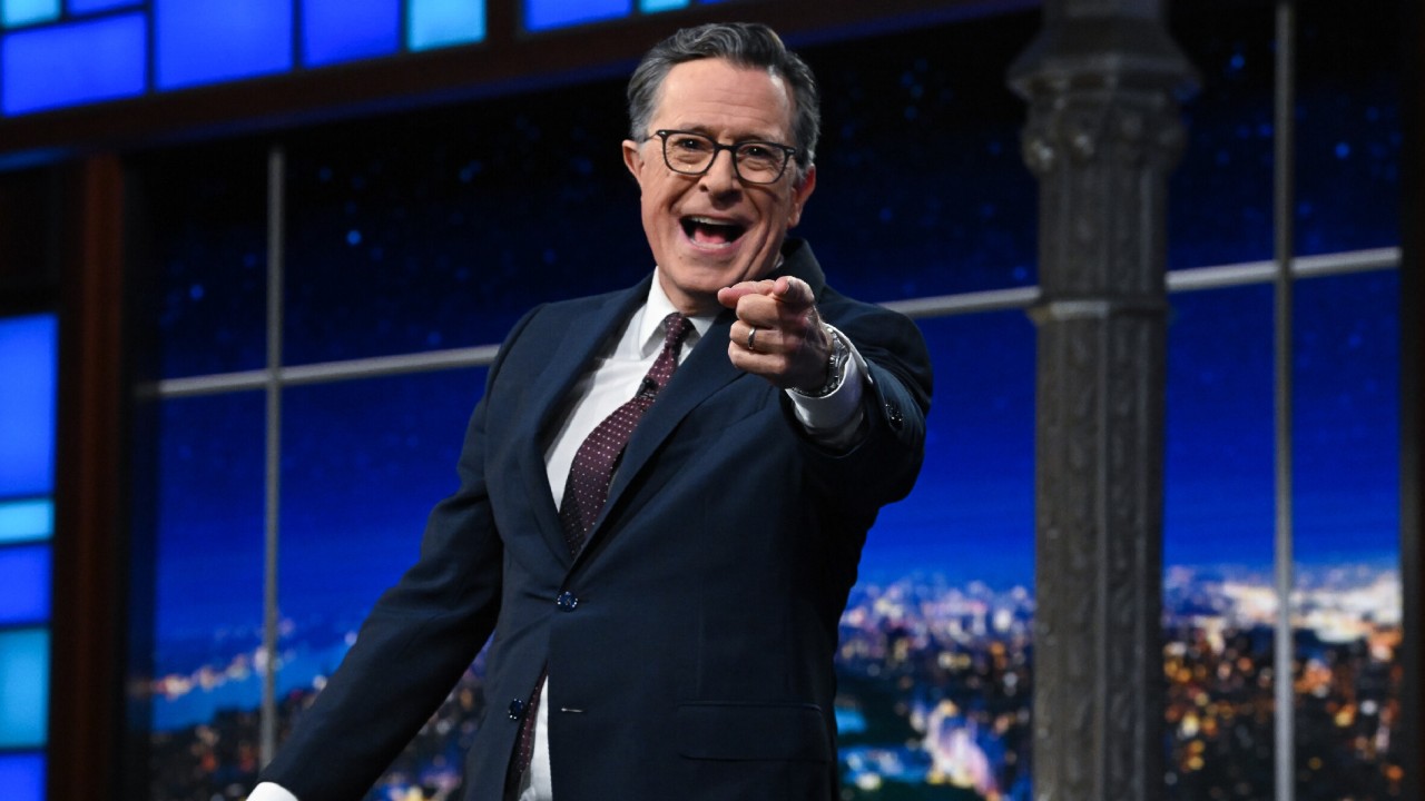 Stephen Colbert smiling and pointing at the camera with his left hand. 