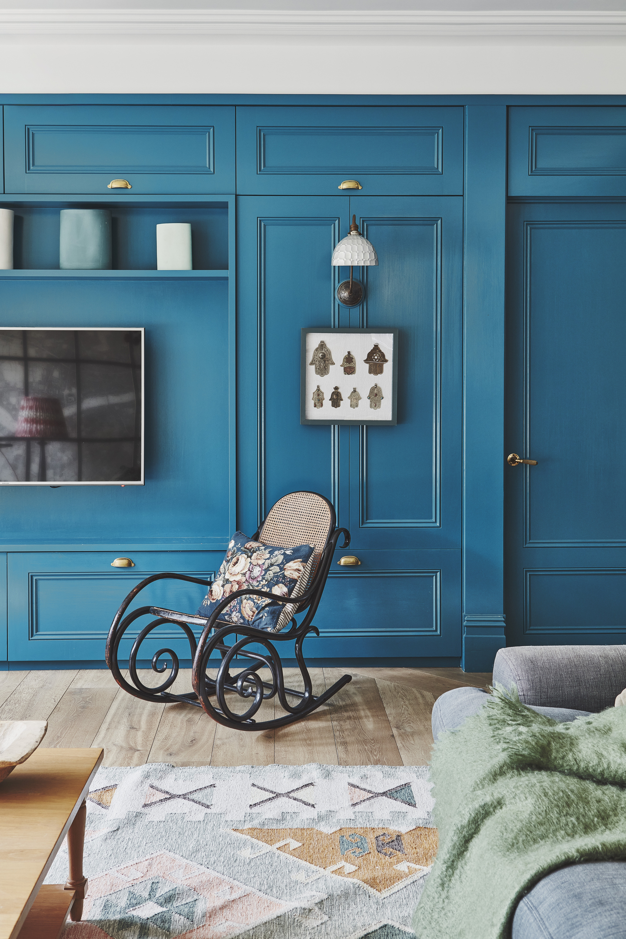 living room with bright blue walls, paneling, rocking chair, patterned rug, grey sofa with green throw and mounted TV