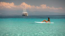 A woman sits on a white surfboard in the clear waters of Indonesia with the Aliikai yacht in the background