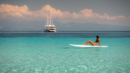 A woman sits on a white surfboard in the clear waters of Indonesia with the Aliikai yacht in the background