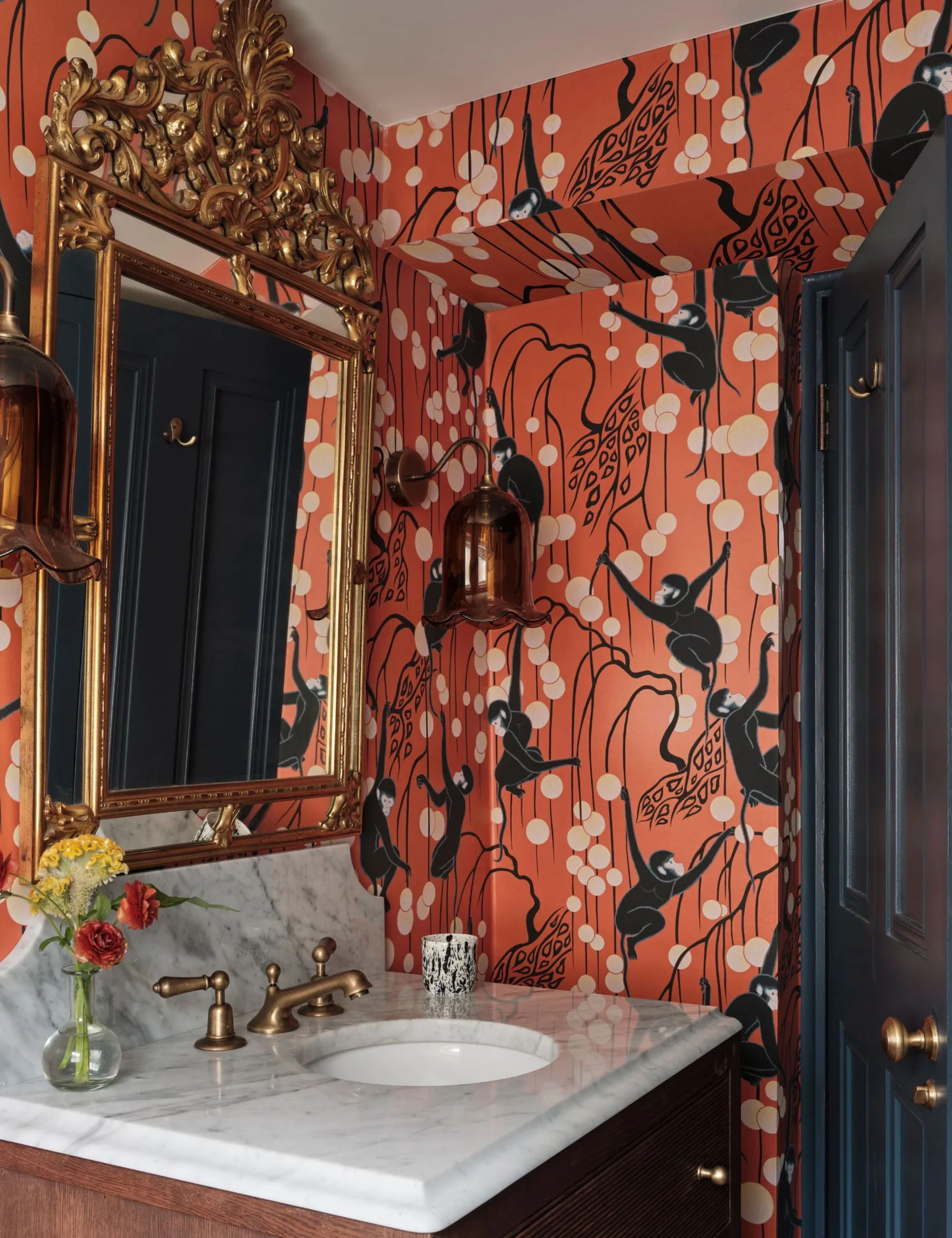 A small guest bathroom decorated with a red patterned wallpaper, a gold mirror, and a marble vanity
