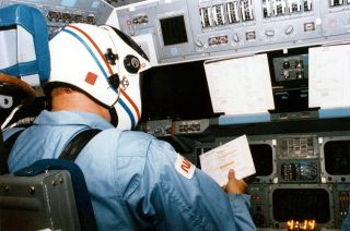 an astronaut in a light-blue flight suite and a white helmet examines note cards on board the space shuttle.