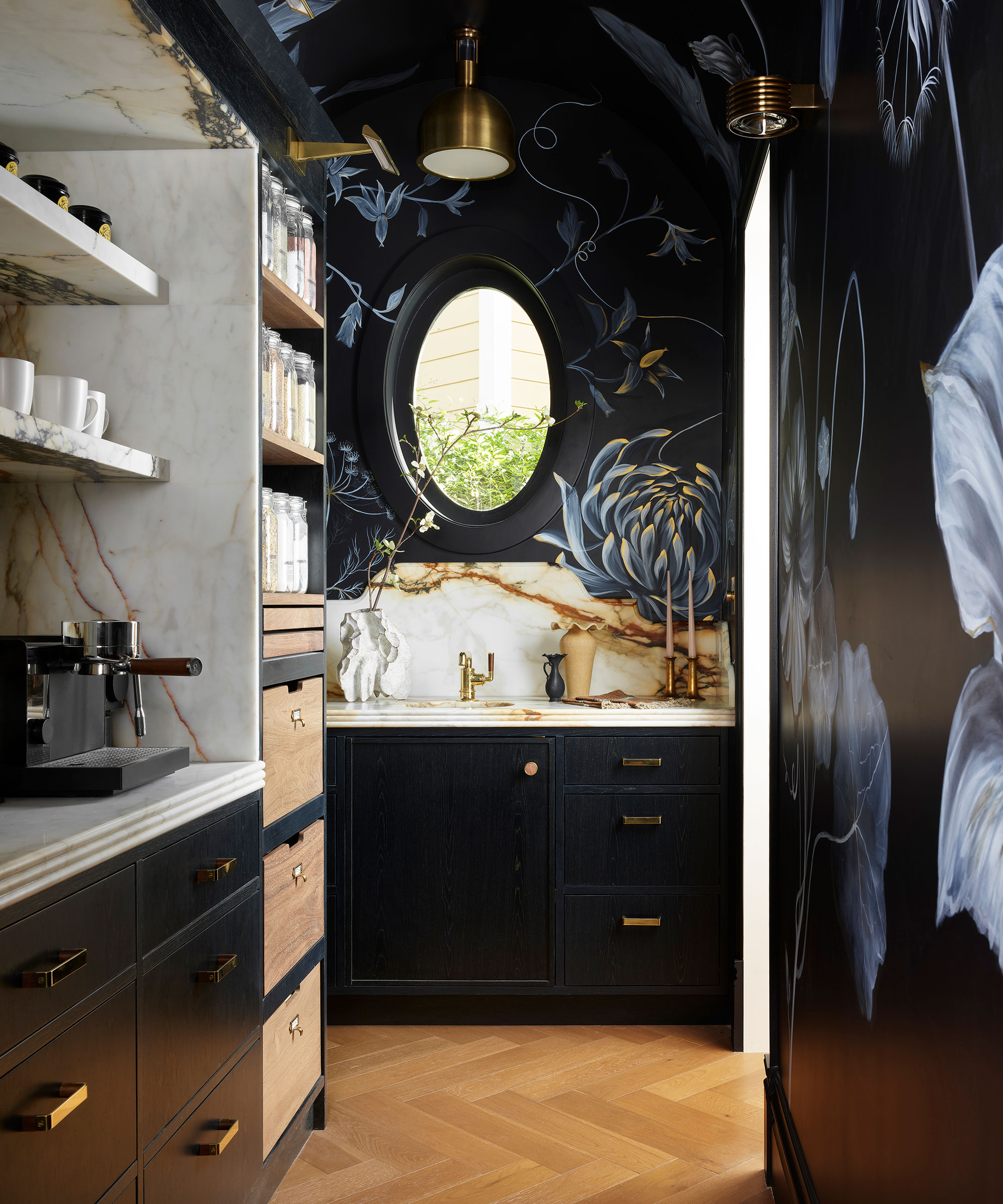 A moody pantry with black floral mural walls, dark cabinetry, and gold hardware. Features include an oval window, marble countertops, herringbone wood floors, and open shelving with glass jars.