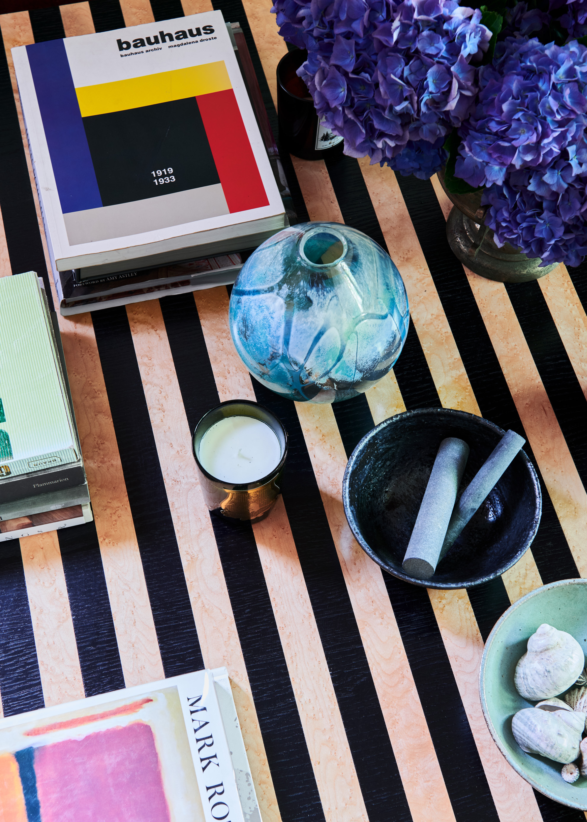 A coffee table vignette featuring a bauhaus book, a vase of purple hydrangeas, a bud vase, a bowl of incense, a bowl of seashells, a scented candle and other books