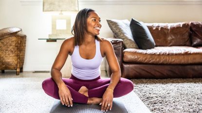 Woman sitting on yoga mat in living room, smiling and looking away from the camera, setting up for the mountain climbers exercise