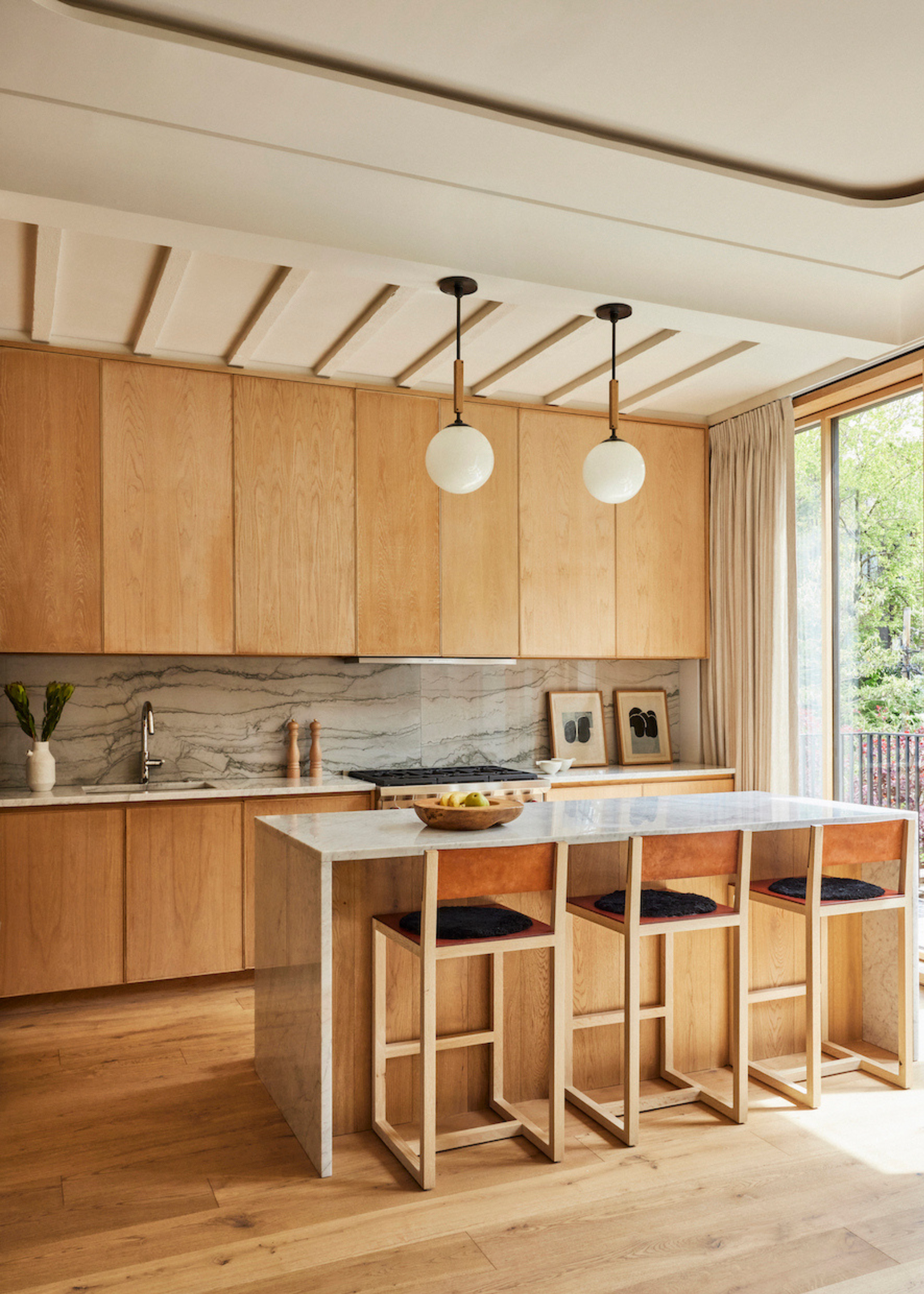 A wood kitchen with pendant ceiling lamps and a stone backsplash. A stone island with a trio of kitchen stools is located in the middle.