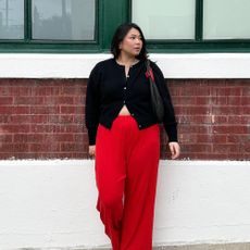 Influencer wears red trousers with a black cardigan, a black woven bag over her shoulder and a pearl necklace.