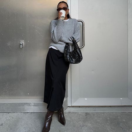 Ingrid wears a gray sweater, black woven bag, black maxi skirt, and black knee-high boots.